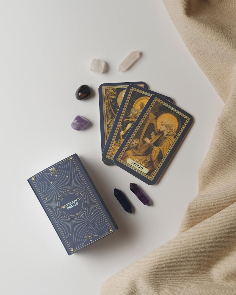 May include: A Mythology Oracle card deck with three cards showing Apollo playing a harp. The deck is navy blue with gold accents, and is surrounded by crystals in various colors. A beige cloth is in the background.