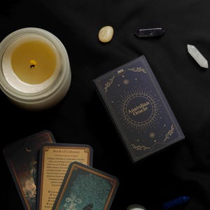 Peut inclure: Vue a&eacute;rienne d'une bougie allum&eacute;e dans un pot en verre, d'un jeu de cartes "Anatolian Oracle" et de plusieurs cristaux sur un tissu noir. Le jeu de cartes est bleu marine avec des lettres dor&eacute;es et un motif circulaire.