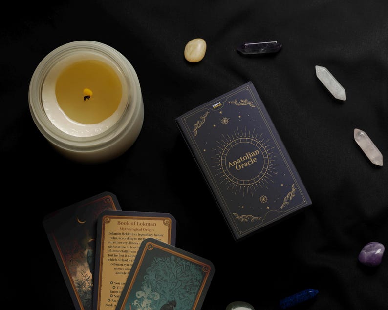 Peut inclure: Vue a&eacute;rienne d'une bougie allum&eacute;e dans un pot en verre, d'un jeu de cartes "Anatolian Oracle" et de plusieurs cristaux sur un tissu noir. Le jeu de cartes est bleu marine avec des lettres dor&eacute;es et un motif circulaire.