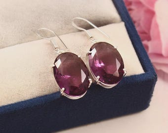 May include: A pair of silver-toned earrings with large, oval, deep purple gemstones. The gemstones are set in a silver metal frame and hang from a simple hook. The earrings are displayed in a blue jewelry box.