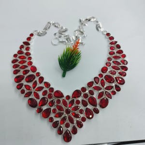 Op de afbeelding: Een zilveren ketting met een slabbetje-ontwerp, versierd met talrijke rode edelstenen in verschillende vormen. De edelstenen zijn dicht op elkaar geplaatst, waardoor een levendig en opvallend sieraad ontstaat. De ketting wordt gepresenteerd op een wit oppervlak.