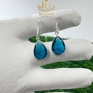 May include: A pair of silver-toned drop earrings featuring teardrop-shaped, faceted, teal-colored gemstones. The earrings are suspended from a coiled wire and are displayed on a white surface. The brand name "BAIDYA JEWELS" is visible in the background.