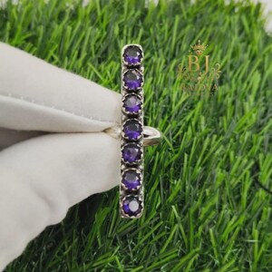 Puede incluir: Un anillo de plata con una barra vertical engastada con siete piedras de amatista moradas, redondas y facetadas. La banda de metal es lisa y pulida, y el engaste muestra un diseño de garras en forma de corona para cada piedra. Logo dorado con texto BJ.