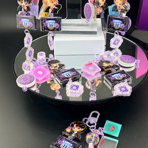 May include: A collection of purple-themed keychains featuring chibi-style character figures, miniature Oreo BTS cookie packages, and small circular charms with various icons. The keychains include light purple plastic clips and translucent beads.