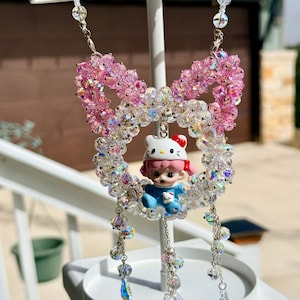May include: A decorative beaded ornament featuring a pink bow and a circular design with a Hello Kitty figurine. The ornament is made of clear and pink beads, with dangling crystal accents. The figurine is wearing a blue outfit and a Hello Kitty hat.