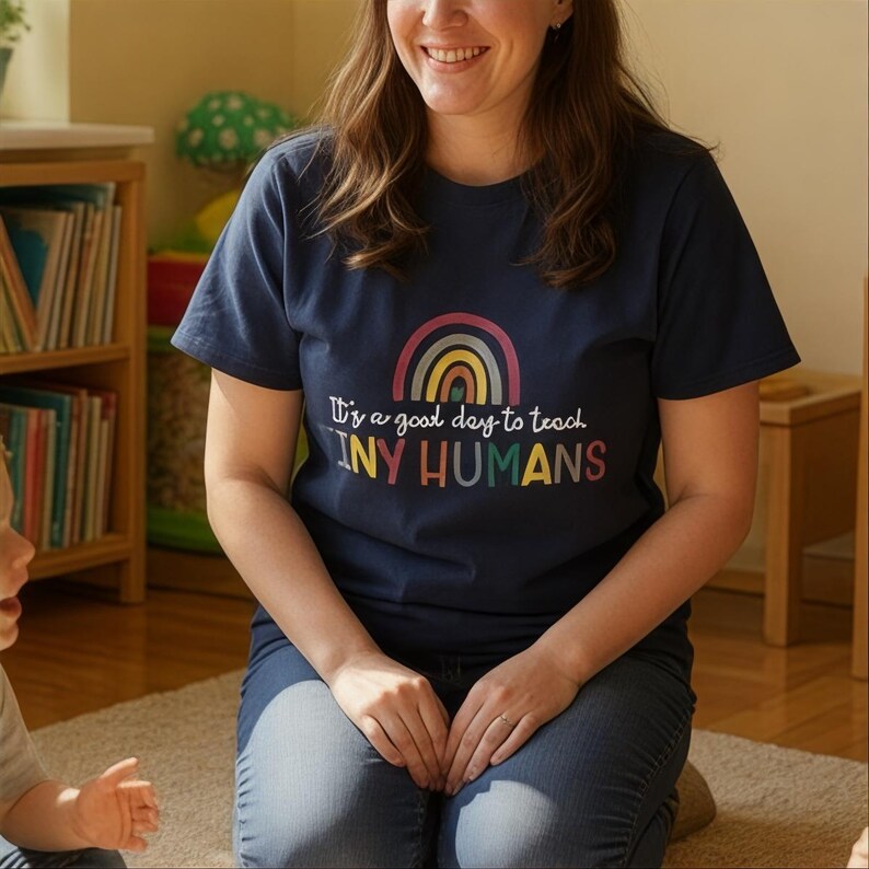 May include: Navy blue t-shirt with a rainbow graphic and the text "It's a good day to teach TINY HUMANS". The shirt is made of a soft, comfortable material.