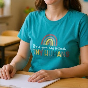 May include: Teal crew neck t-shirt with a rainbow graphic and the text "It's a good day to teach TINY HUMANS". The shirt is made of a soft material.