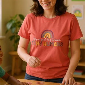May include: A coral-colored t-shirt with a rainbow graphic and the text "It's a good day to teach TINY HUMANS." The shirt has short sleeves and a crew neck. The design features a rainbow in shades of purple, yellow, and blue.