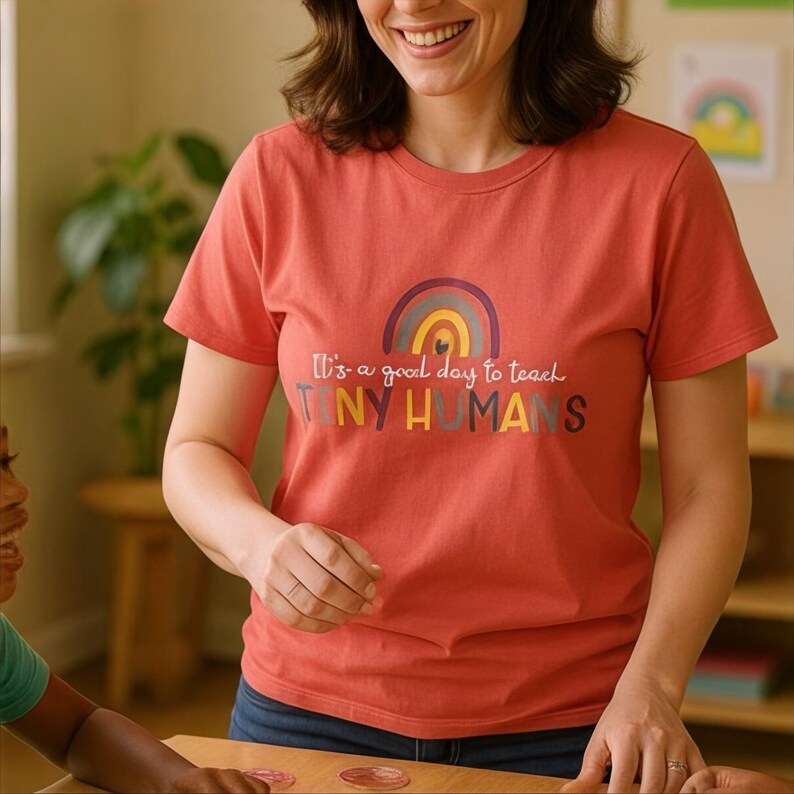 May include: A coral-colored t-shirt with a rainbow graphic and the text "It's a good day to teach TINY HUMANS." The shirt has short sleeves and a crew neck. The design features a rainbow in shades of purple, yellow, and blue.