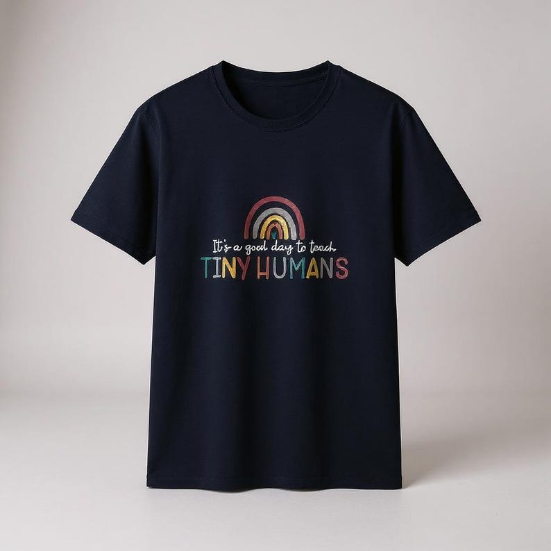 May include: Navy blue t-shirt with a rainbow graphic and the text "It's a good day to teach TINY HUMANS" in colorful letters. The shirt has short sleeves and a crew neck.