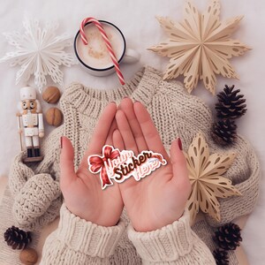 May include: A sticker with a red bow and the text "Your Sticker Here" is held in open hands. The sticker is surrounded by a cream-colored sweater, paper snowflakes, pine cones, a mug of coffee with a candy cane, and a wooden nutcracker.