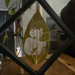 Op de afbeelding: Een ingelijst kunstwerk met een portret geëtst op een blad. Het blad, met zijn ingewikkelde nerven, is ingesloten in een zwart, ruitvormig frame. Het kunstwerk wordt tentoongesteld op een houten oppervlak, met groen op de achtergrond.