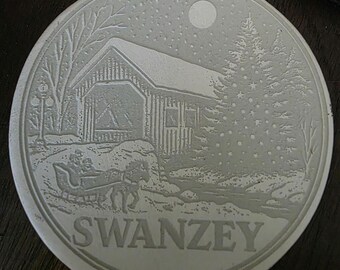 May include: A circular, off-white Christmas ornament featuring a winter landscape. The design depicts a covered bridge, a horse-drawn sleigh, a Christmas tree, and the word "SWANZEY". A full moon and falling snow are also visible.