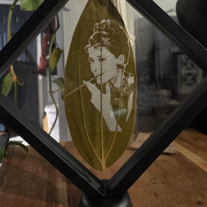 Op de afbeelding: Een ingelijst bladkunstwerk met een portret van een vrouw met een sigarettenhouder. De afbeelding is een witte ets op een groen blad, ingesloten in een zwart, ruitvormig frame. Het frame wordt ondersteund door een zwarte voet, weergegeven op een houten oppervlak.