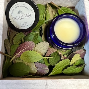 May include: A wooden box filled with green and purple leaves, and two small jars. One jar is black with white text that reads "Midnight Bloom Apothecary Lyreleaf Sage Salve." The other jar is blue and contains a yellow salve.