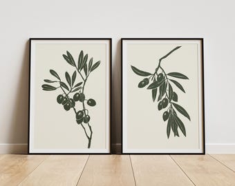 May include: Two framed art prints featuring olive branch illustrations. The artwork depicts dark green olive branches with leaves and olives against a cream background. The frames are black, and the prints are displayed on a light wood floor.