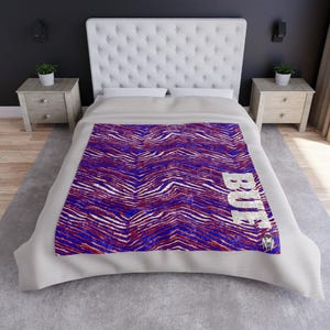May include: A rectangular throw blanket featuring a vibrant red, white, and blue zebra-striped pattern. The word "BUE" is printed in large, distressed white capital letters along the right side, accompanied by a small graphic of a buffalo head.