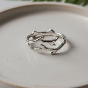 Op de afbeelding: Een zilverkleurige ring met een takachtig ontwerp. De ring ligt op een witte keramische plaat, met een wazig groen blad op de achtergrond. Het ontwerp van de ring heeft meerdere gebogen takken. Deze ring is een sieraad.