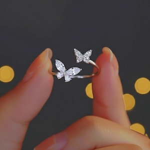May include: A silver ring with two butterfly designs, each embellished with clear, sparkling gemstones. The ring's band is slender, and the butterflies are arranged to give the impression of fluttering around the finger. The ring is held against a blurred background.