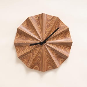 May include: A wooden wall clock with a distinctive geometric design. The clock face is made of layered wood, displaying a natural grain pattern. Black clock hands show the time. The clock is mounted on a plain, off-white wall.