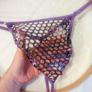 May include: A purple and black thong with a mesh design. The thong features a shiny, glittery fabric and thin straps. The garment is held up by a hand, with a wooden embroidery hoop in the background.
