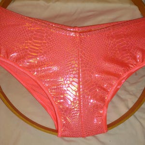 May include: High-waisted underwear in a vibrant neon coral colour featuring a reflective, iridescent snakeskin-like scale pattern across the entire surface. The garment has a smooth, shiny finish and a central vertical seam running down the front.