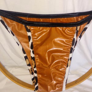 May include: A pair of brown and black underwear with a shiny, wet look. The sides feature a leopard print pattern, and the edges are trimmed with black fabric. The underwear is displayed on a wooden hoop.