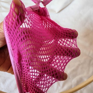 May include: A bright pink lingerie set featuring a fishnet design. The set includes a halter top with adjustable straps and a matching bottom. The fabric is a vibrant fuchsia color, and the design is open and airy.