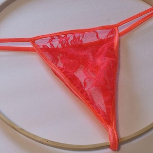 May include: A bright orange lace thong with a delicate floral pattern. The thong is displayed on a wooden embroidery hoop.