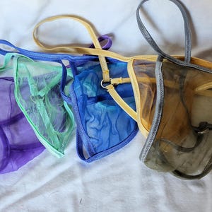 Puede incluir: Una colección de tangas transparentes en varios colores, incluyendo morado, verde, azul, amarillo y gris. La lencería presenta delicadas correas y una tela de malla transparente, creando una apariencia ligera y aireada.