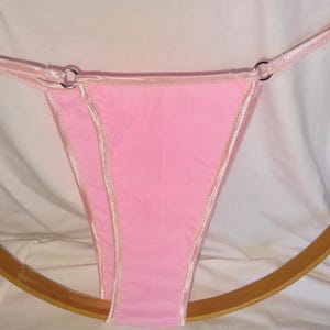 May include: A light pink thong-style underwear with a satin trim and straps. The straps are attached to the main body with silver metal rings. The underwear is displayed on a wooden hoop.
