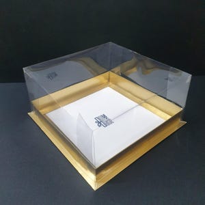 Könnte beinhalten: Eine transparente, quadratische Kunststoffbox mit goldfarbenem Boden und Zierleisten. Die Box hat einen weißen Innenraum und einen transparenten Deckel. Die Box ist für Verpackung und Präsentation konzipiert.