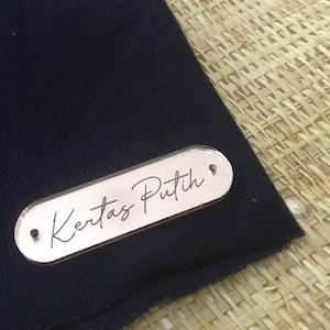 Puede incluir: Una etiqueta rectangular de oro rosa con las palabras "Kertas Putih" en escritura cursiva. La etiqueta está sobre una tela azul marino oscuro. La etiqueta tiene dos pequeños agujeros en cada extremo.