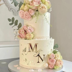 Può includere: Una torta nuziale a due piani con glassa color avorio, ornata di rose rosa e gialle. La torta presenta le iniziali dorate "M & N" e dettagli di foglie d'argento. La torta è su un supporto argentato.