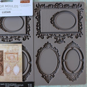 May include: A gray silicone mold with six different ornate oval frame designs. The mold is 5 inches by 8 inches and is food safe. The text "Decor Moulds" and "Lucian" is printed on the packaging.