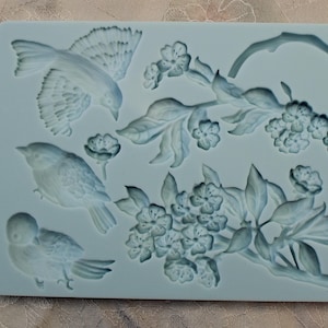 Puede incluir: Un molde de silicona azul con un diseño de tres pájaros y una rama con flores. El molde está sobre una superficie beige claro. El texto "Ir-design" es visible en el molde.