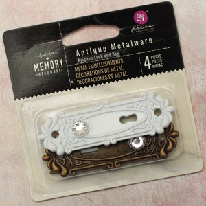 May include: A package of four antique metalware embellishments. The set includes two Avignon lock and key designs. One is white with a crystal accent, and the other is bronze-colored. The package is labeled "Memory Hardware."