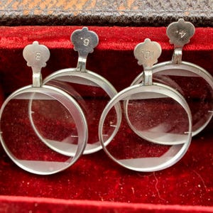 May include: Four antique silver-rimmed spectacles with clear lenses, each with a small, numbered handle. The spectacles are presented in a red velvet-lined case, with the numbers 4, 8, 9, and 10 visible on the handles.