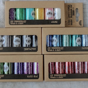 May include: Six sets of glitter in various colours, each set is packaged in a brown cardboard box with a clear window showing the glitter. The boxes are labelled with the words "Glitter Set" and the name of the colour set. The colour sets are: Abloom, Ebony & Ivory, Caribbean Seas, Mardi Gras, and Crimson.