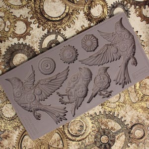 May include: A grey silicone mould featuring detailed bird designs with mechanical gear accents. The mould includes birds in various poses, along with circular gear elements. The background showcases a pattern of golden gears.