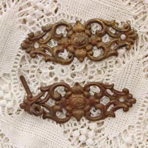 May include: Two antique bronze-colored drawer pulls with ornate, symmetrical designs. Each pull features a central oval shape surrounded by swirling, floral-like patterns. One pull has a screw attached.