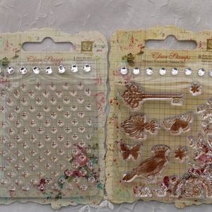 May include: Two clear stamp sets with a vintage floral design. The first set features a grid of small circles and crosses. The second set features a key, a bird, a butterfly, and other floral designs. The text on the packaging reads "Clear Stamps".