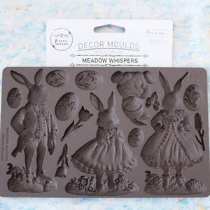 以下が含まれることがあります： イースターをテーマにしたデザインのグレーのシリコンモールド。ヴィンテージの服を着た詳細なウサギのフィギュア、イースターエッグ、花のアクセントが含まれています。「Decor Moulds」と「Meadow Whispers」と書かれたパッケージ。