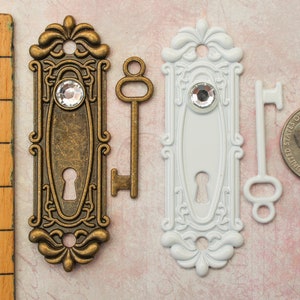 May include: Two decorative door plates, one bronze and one white, each with a crystal accent and keyhole. Two keys, one bronze and one white, are also present. A wooden ruler shows measurements in inches.