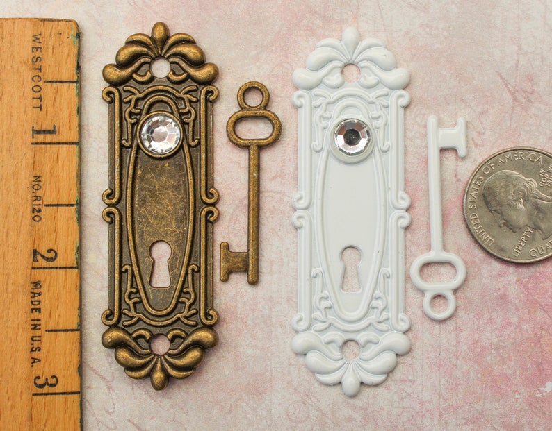 May include: Two decorative door plates, one bronze and one white, each with a crystal accent and keyhole. Two keys, one bronze and one white, are also present. A wooden ruler shows measurements in inches.