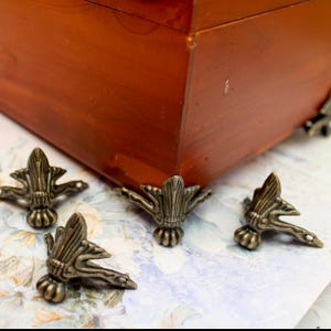 Puede incluir: Patas de muebles ornamentadas de metal color bronce. Las patas tienen un diseño detallado y estilizado con una base redondeada y elementos que se curvan hacia afuera. Se colocan cerca de la esquina de una caja de madera con acabado marrón rojizo.