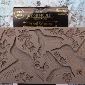 May include: A rectangular silicone decor mould in a taupe colour featuring embossed designs of three hummingbirds, several cherry blossom branches, and two small crowns. The black header card displays the text DECOR MOULDS BLOOM & FLUTTER.