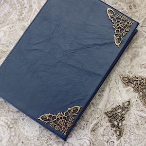 May include: A navy blue leather-bound journal with ornate bronze corner protectors. Two additional bronze corner pieces are placed nearby. The journal is on a patterned, off-white surface.