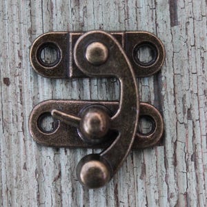 Op de afbeelding: Een bronzen metalen sluiting met een gebogen grendel en twee rechthoekige platen, elk met twee cirkelvormige gaten. De sluiting is ontworpen voor het vastzetten van kleine dozen of decoratieve items. De achtergrond is een verweerd, lichtblauw houten oppervlak.