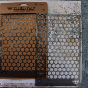 May include: Two stencils with a honeycomb pattern. The stencils are made of a silver-colored material. The packaging is brown with the words "ELEMENTALS" and "Honeycomb" printed on it. The stencils are designed for crafting and mixed media projects.